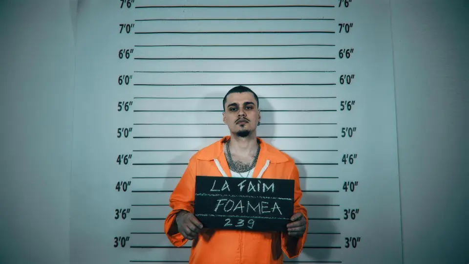 Mugshotul oficial al lui Foamea: uniformă portocalie, placa LA FAIM / FOAMEA / 239, perete cu linii de înălțime